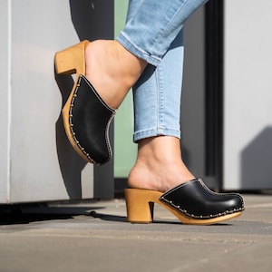 Puede incluir: Zuecos de cuero negro con tacón de madera y tachuelas doradas. Los zuecos tienen la punta cerrada y una forma redondeada.