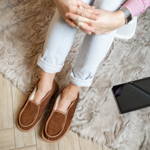 May include: A pair of brown suede slippers with a shearling lining. The slippers are worn by a person sitting on a fluffy rug.