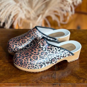 Puede incluir: Un par de zuecos con estampado de leopardo. Los zuecos tienen suela de madera, plantilla blanca y ribete negro. Los zapatos est&aacute;n sobre una superficie de madera.