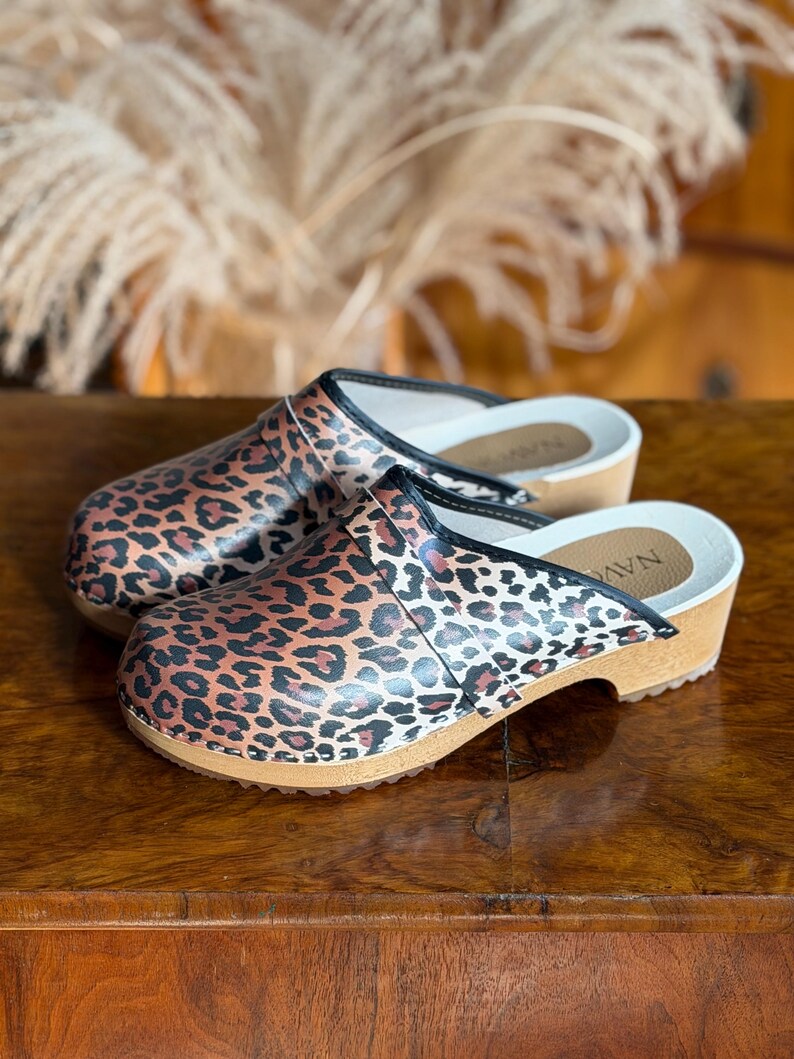 Puede incluir: Un par de zuecos con estampado de leopardo. Los zuecos tienen suela de madera, plantilla blanca y ribete negro. Los zapatos est&aacute;n sobre una superficie de madera.