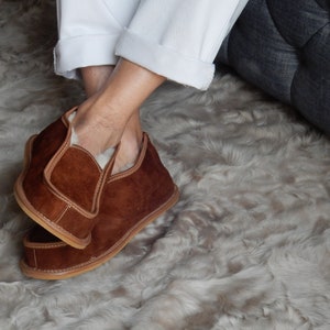 May include: Brown suede slippers with a shearling lining. The slippers are worn on a fluffy white rug.