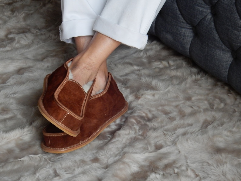 May include: Brown suede slippers with a shearling lining. The slippers are worn on a fluffy white rug.