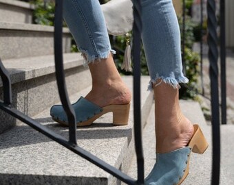 May include: A person wearing blue denim jeans and light blue suede clogs with wooden heels. The clogs have a pointed toe and are decorated with small metal studs.