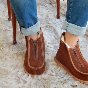 May include: Brown suede slippers with a zipper up the front and a white fur lining. The slippers are worn by a person sitting on a chair with their legs crossed.
