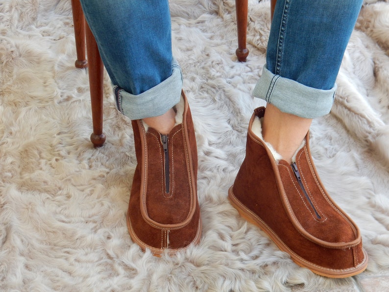 May include: Brown suede slippers with a zipper up the front and a white fur lining. The slippers are worn by a person sitting on a chair with their legs crossed.