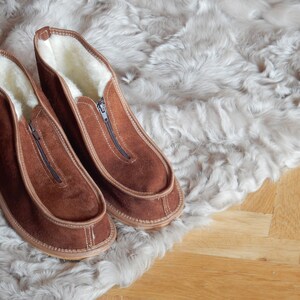 May include: A pair of brown suede slippers with a zipper on the front. The slippers are lined with white fleece and are sitting on a white faux fur rug.