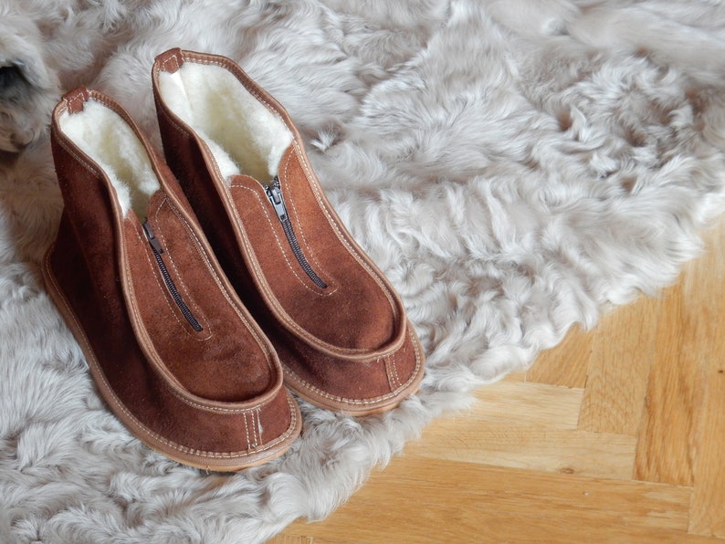 May include: A pair of brown suede slippers with a zipper on the front. The slippers are lined with white fleece and are sitting on a white faux fur rug.