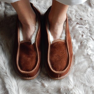 May include: A pair of brown suede slippers with a white sheepskin lining. The slippers are worn on a white fluffy rug.