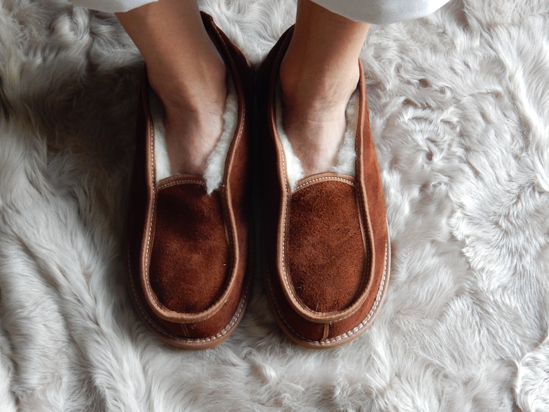 May include: A pair of brown suede slippers with a white sheepskin lining. The slippers are worn on a white fluffy rug.