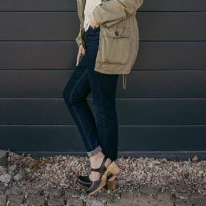 Può includere: Una persona indossa jeans blu scuro, zoccoli neri con tacco in legno e una giacca verde oliva. Gli zoccoli hanno un cinturino sulla parte superiore del piede e un tacco in legno. La giacca ha tasche e un cordoncino.