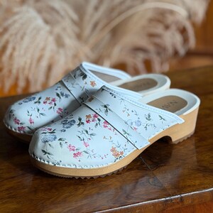 Puede incluir: Un par de zuecos blancos con un estampado floral con flores rosas, azules y naranjas. Los zuecos tienen una suela de madera y una correa de cuero blanca. Los zuecos se exhiben sobre una superficie de madera.