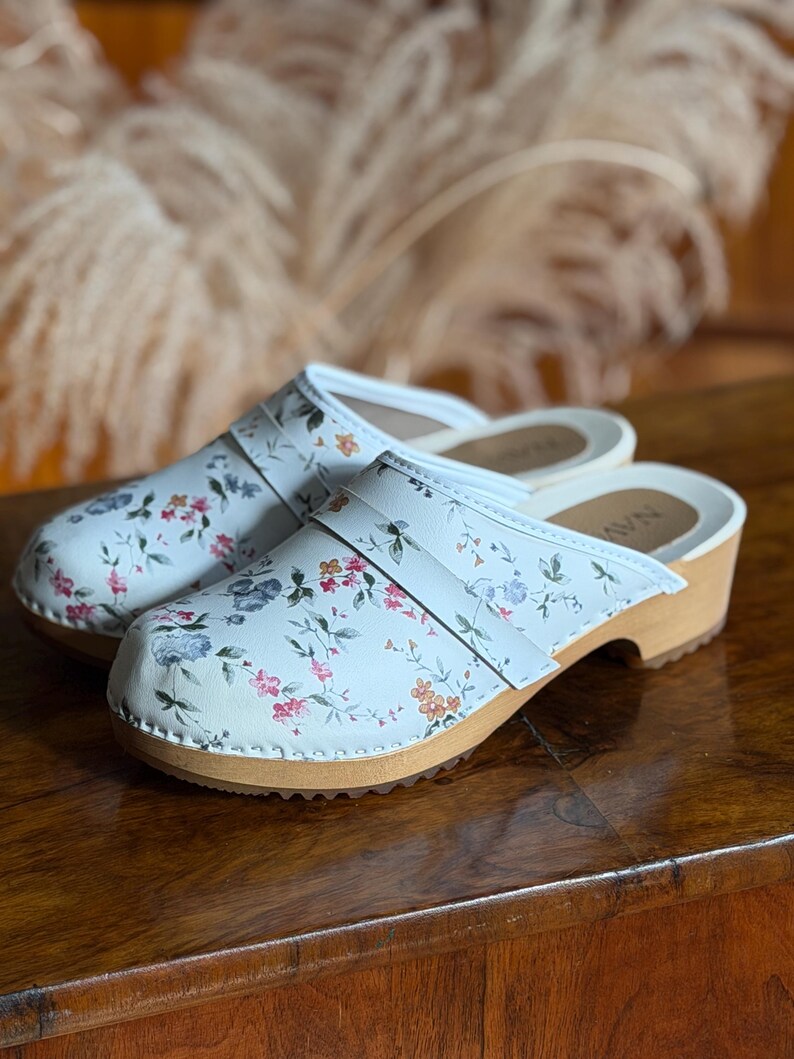 Puede incluir: Un par de zuecos blancos con un estampado floral con flores rosas, azules y naranjas. Los zuecos tienen una suela de madera y una correa de cuero blanca. Los zuecos se exhiben sobre una superficie de madera.