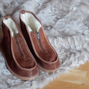 May include: A pair of brown suede slippers with a zipper on the front. The slippers are lined with white sheepskin and have a brown leather sole.
