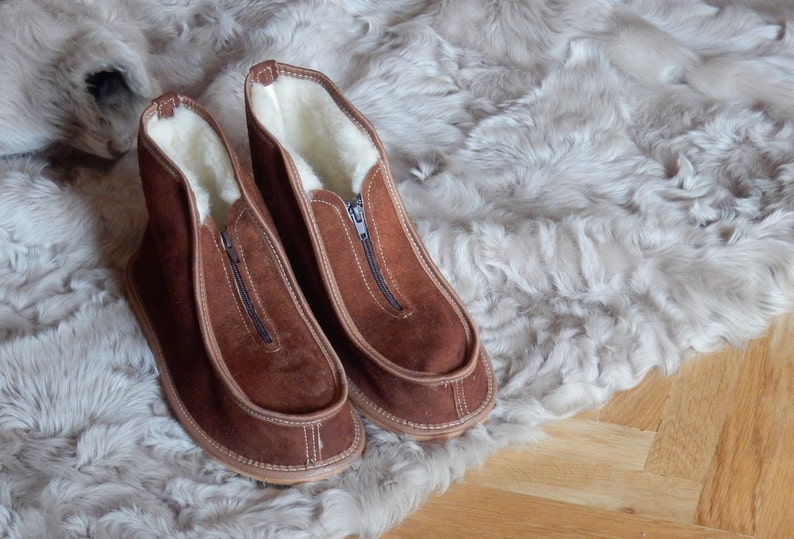 May include: A pair of brown suede slippers with a zipper on the front. The slippers are lined with white sheepskin and have a brown leather sole.