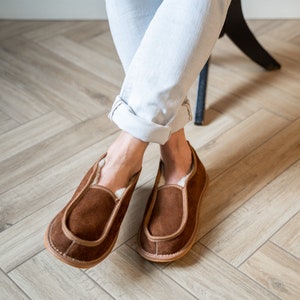 May include: A pair of brown suede slippers with a white sheepskin lining. The slippers have a simple design with a rounded toe and a flat sole.