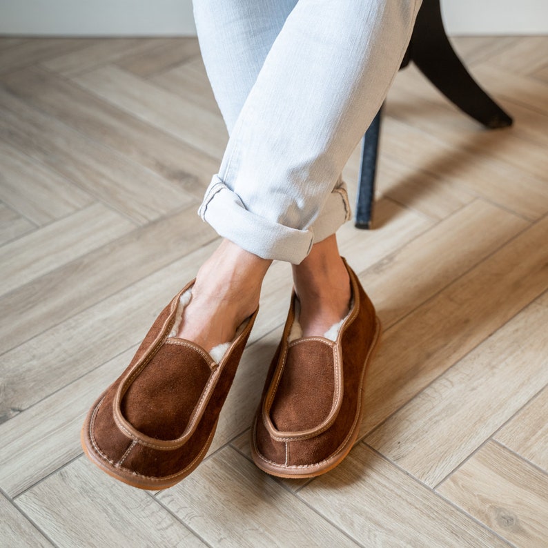 May include: A pair of brown suede slippers with a white sheepskin lining. The slippers have a simple design with a rounded toe and a flat sole.
