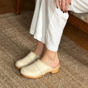 Puede incluir: Zuecos de cuero color crema con suela de madera. Los zuecos tienen una correa en la parte superior. Los zuecos los lleva una persona con pantalones blancos. Los zuecos est&aacute;n sobre una alfombra tejida.