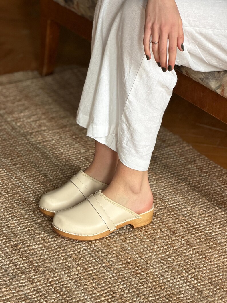 Puede incluir: Zuecos de cuero color crema con suela de madera. Los zuecos tienen una correa en la parte superior. Los zuecos los lleva una persona con pantalones blancos. Los zuecos est&aacute;n sobre una alfombra tejida.