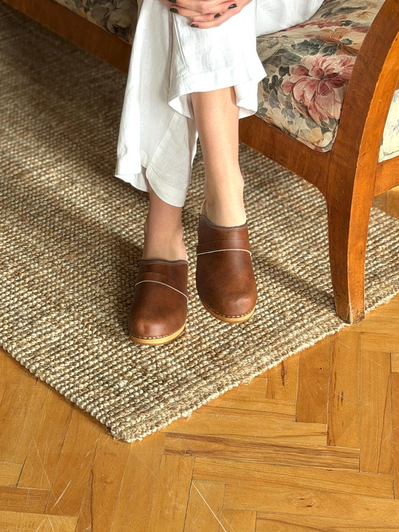Puede incluir: Zuecos de cuero marr&oacute;n con suela de color claro y una fina l&iacute;nea blanca. Los zuecos se usan sobre una alfombra de yute tejida, con una silla de madera y una persona que usa pantalones blancos en el fondo.