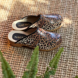 Puede incluir: Un par de zuecos con estampado de leopardo. Los zuecos tienen suela de madera y ribete negro. El nombre de la marca "NAVONA" es visible en la plantilla. Los zuecos est&aacute;n sobre una superficie tejida marr&oacute;n.