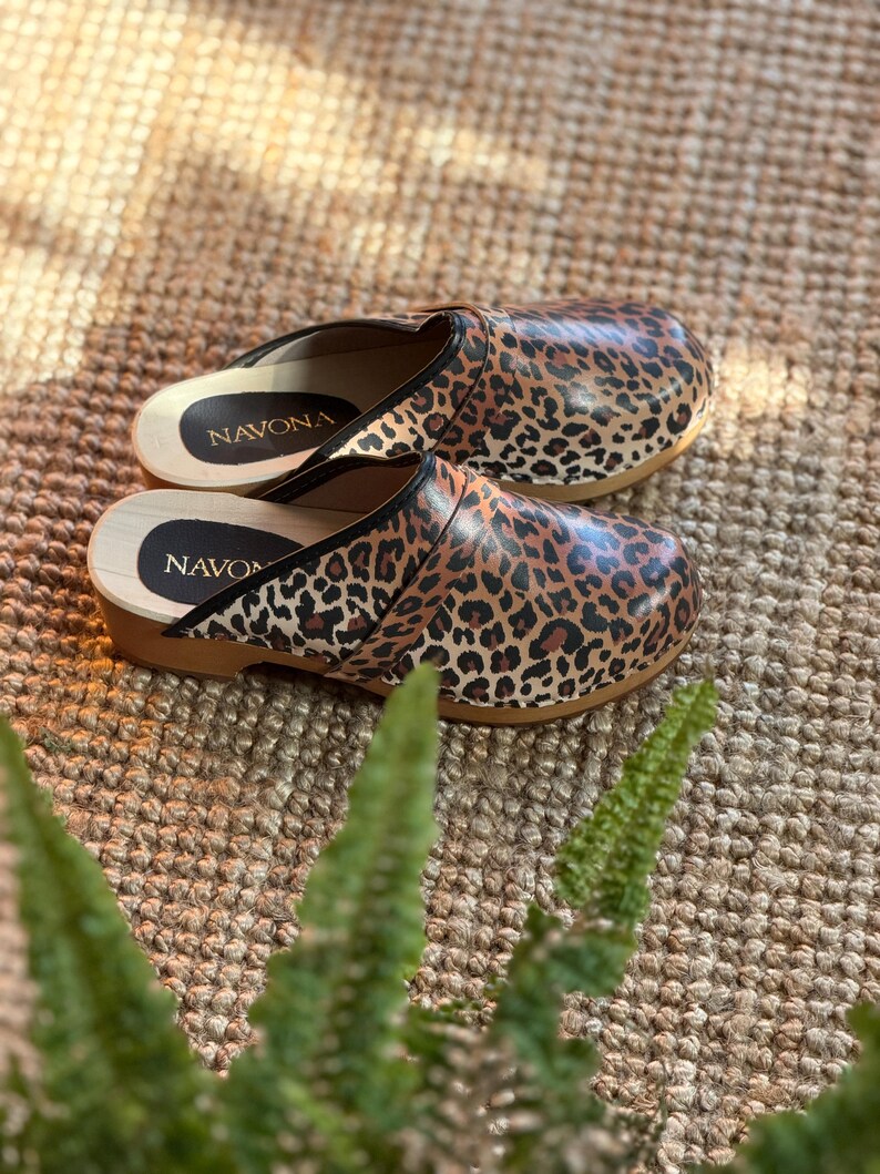 Puede incluir: Un par de zuecos con estampado de leopardo. Los zuecos tienen suela de madera y ribete negro. El nombre de la marca "NAVONA" es visible en la plantilla. Los zuecos est&aacute;n sobre una superficie tejida marr&oacute;n.