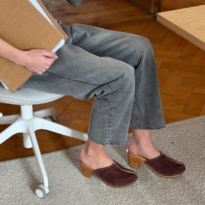 May include: Brown suede clogs with wooden heels. The clogs feature a rounded toe and small, gold-coloured studs along the sole. The heels are approximately 7.6 cm high. These clogs are a classic style of footwear, suitable for various occasions.
