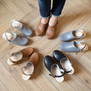 Może przedstawiać: Różne drewniaki skórzane w odcieniach szarości, brązu i czerni ułożone na drewnianej podłodze. Drewniaki mają drewniane podeszwy i skórzane paski z klamrami. Jedna para brązowych drewniaków jest noszona.