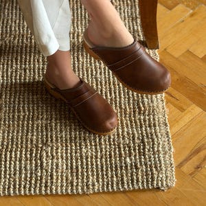 Puede incluir: Zuecos de cuero marr&oacute;n con suela de madera. Los zuecos tienen un dise&ntilde;o de punta cerrada y dos correas en la parte superior. Los zapatos se usan sobre una alfombra de yute tejida. Los zuecos son de estilo cl&aacute;sico.