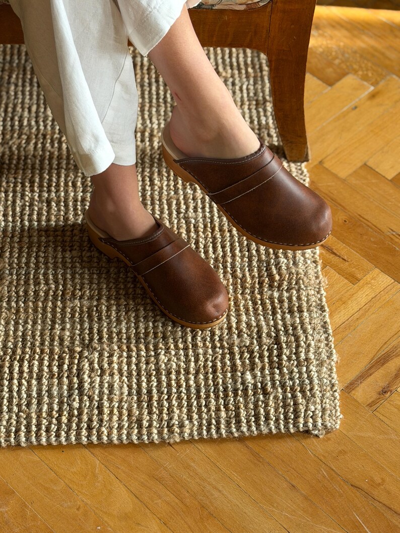 Puede incluir: Zuecos de cuero marr&oacute;n con suela de madera. Los zuecos tienen un dise&ntilde;o de punta cerrada y dos correas en la parte superior. Los zapatos se usan sobre una alfombra de yute tejida. Los zuecos son de estilo cl&aacute;sico.