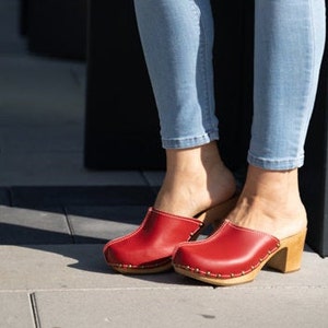 Puede incluir: Zuecos de cuero rojo con tacón de madera y puntera cerrada. Los zuecos los lleva una persona que lleva pantalones vaqueros azules.
