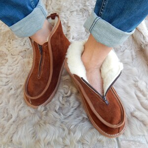 May include: Brown suede slippers with a zipper and white faux fur lining. The slippers are worn on a light brown fuzzy rug.