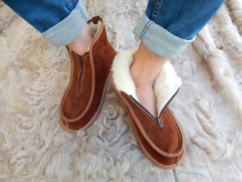 May include: Brown suede slippers with a zipper and white faux fur lining. The slippers are worn on a light brown fuzzy rug.