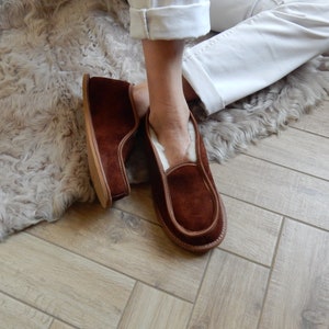 May include: A pair of brown suede slippers with a white sheepskin lining. The slippers are worn by a person sitting on a fluffy rug.
