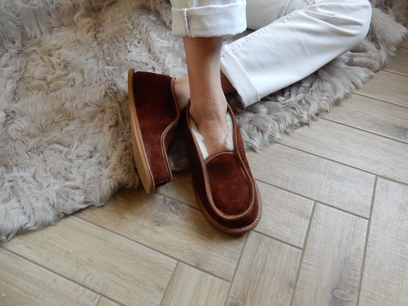 May include: A pair of brown suede slippers with a white sheepskin lining. The slippers are worn by a person sitting on a fluffy rug.