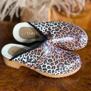 Puede incluir: Un par de zuecos con estampado de leopardo. Los zuecos tienen suela de madera y ribete negro. El interior es de color beige claro. La marca "NACHA" es visible en el interior de un zueco.