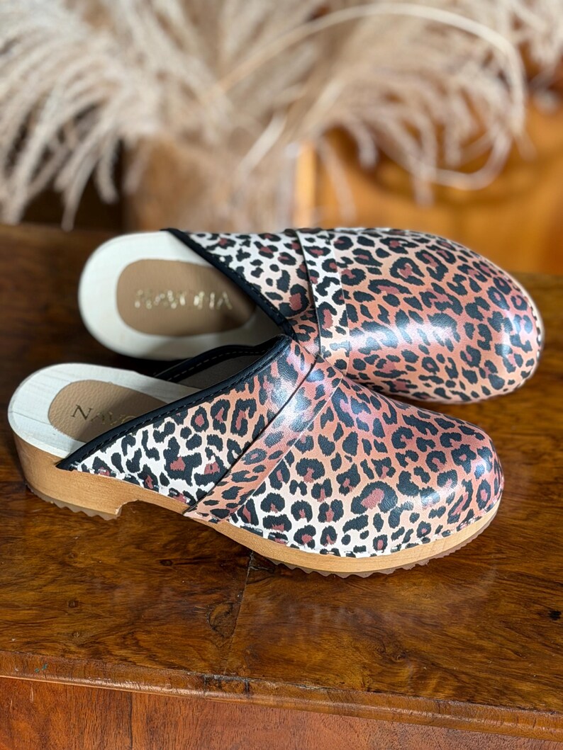 Puede incluir: Un par de zuecos con estampado de leopardo. Los zuecos tienen suela de madera y ribete negro. El interior es de color beige claro. La marca "NACHA" es visible en el interior de un zueco.