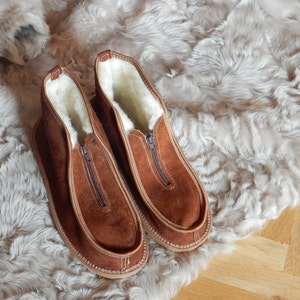 May include: A pair of brown suede slippers with a zipper closure and a white sheepskin lining. The slippers are on a white faux fur background.