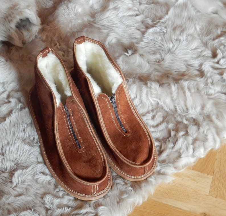 May include: A pair of brown suede slippers with a zipper closure and a white sheepskin lining. The slippers are on a white faux fur background.