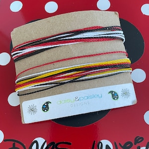 May include: A close-up of a brown paper card with red, white, black, and yellow string wrapped around it. The card has a small white sticker with the text "daisy & paisley designs" on it. The card is sitting on a red surface with white polka dots. The text "DAPS UNTIL DISNEY" is written in black on the red surface.