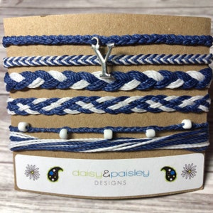 Könnte beinhalten: Ein Set aus sieben blauen und weißen Freundschaftsarmbändern mit einem silbernen Anhänger in Form des Buchstabens Y. Die Armbänder werden auf einer braunen Karte mit dem Text "daisy&paisley DESIGNS" präsentiert.