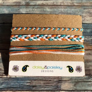 Puede incluir: Tres pulseras de la amistad de color turquesa, blanco y naranja sobre una tarjeta marrón con el texto "daisy&paisley DESIGNS".