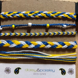 May include: Five friendship bracelets in blue, white, and yellow colors. The bracelets are made with braided string and some have small beads. The bracelets are displayed on a brown surface with a business card that reads "daisy&paisley DESIGNS".