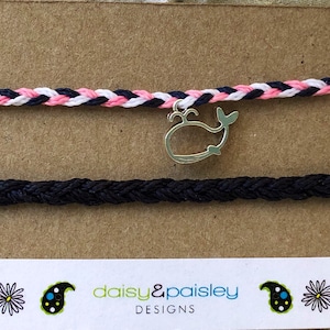 May include: Two friendship bracelets with a whale charm. The top bracelet is white, pink, and black. The bottom bracelet is black. The charm is silver and has a whale design. The bracelets are on a brown background with the text "daisy&paisley DESIGNS".