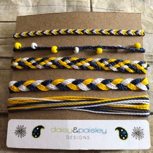 Puede incluir: Cinco pulseras hechas a mano en tonos azul marino, amarillo y blanco. Las pulseras presentan diseños trenzados y con cuentas. La parte inferior de la imagen muestra una etiqueta con el texto "daisy & paisley DESIGNS".