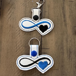 May include: Two white keychains with an infinity symbol and a heart design. One keychain has a black heart and blue infinity symbol, the other has a blue heart and black infinity symbol. Each keychain has a different clasp.