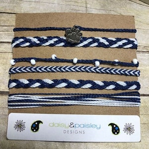 May include: A set of six navy blue and white braided friendship bracelets with white beads and a silver paw print charm. The bracelets are displayed on a brown card with the text "daisy & paisley DESIGNS".