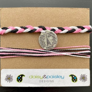 Puede incluir: Un brazalete trenzado negro, blanco y rosa con un dije plateado que presenta la Torre Eiffel y el texto "La tour Eiffel est la tour symbole de Paris" y "PARIS". El brazalete está empaquetado en una tarjeta marrón con una etiqueta blanca que dice "daisy&paisley DESIGNS".