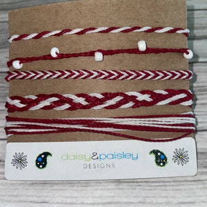Op de afbeelding: Een verzameling van zes handgemaakte armbanden in rood en wit, gepresenteerd op een bruine kaart. De armbanden hebben verschillende gevlochten en geknoopte ontwerpen, sommige met kleine witte kralen. De kaart toont de tekst "daisy & paisley DESIGNS".