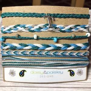 Puede incluir: Un conjunto de siete pulseras de la amistad en tonos de verde azulado, blanco y gris. Las pulseras están hechas de cuerda trenzada y tienen pequeñas cuentas. El conjunto incluye un dije con la letra "T". La tarjeta dice "daisy&paisley DESIGNS".
