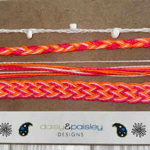 Könnte beinhalten: Satz von fünf Freundschaftsarmbändern in Rosa, Orange und Weiß. Die Armbänder werden auf einer braunen Karte mit dem Text "daisy & paisley DESIGNS" präsentiert.